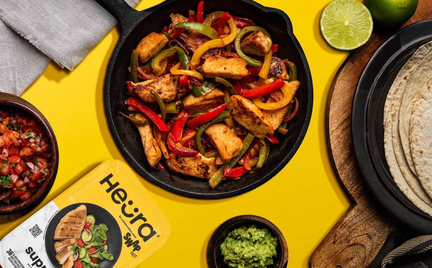 Photo shows a chopped up chicken fillet from Heura and Swap in a cast iron pan along with peppers and onions, against a yellow background, and alongside the product's new packaging