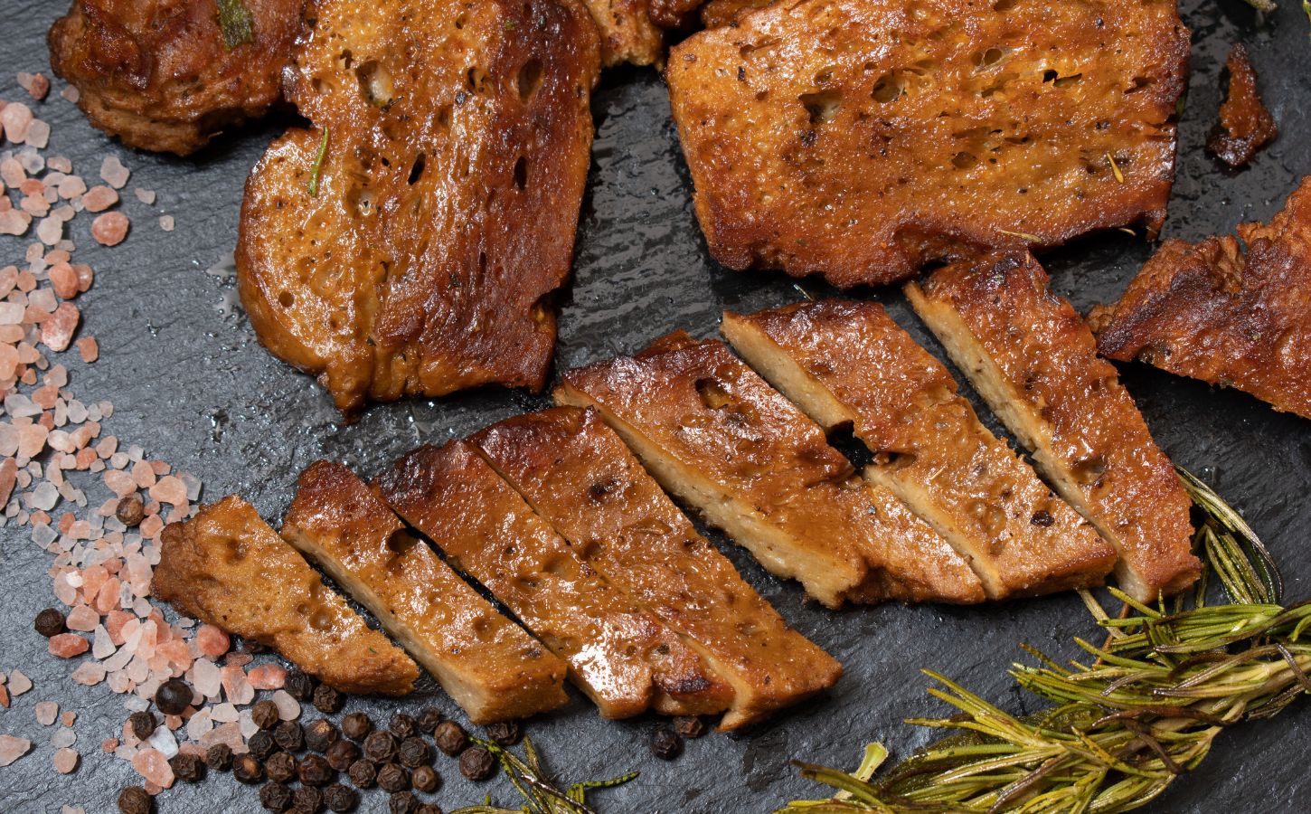 Photo shows several seitan steaks on a slate alongside rock salt, pepper, and rosemary