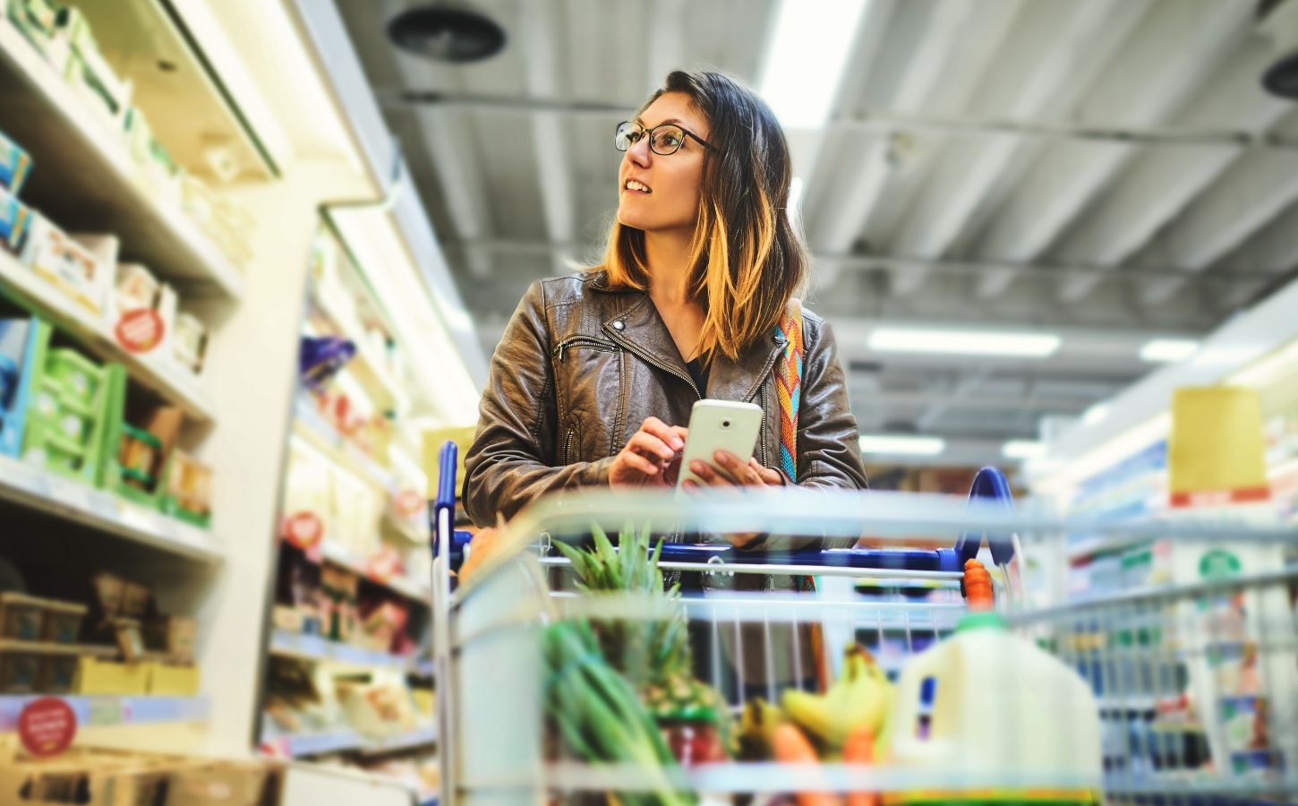 Photo shows a young woman shopping in a supermarket while holding her phone and pushing a trolley